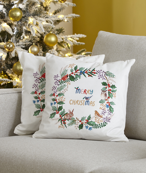 Two festive Christmas wreath cushions on a sofa with a Christmas tree behind them.  